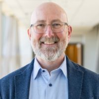Portrait of a man with glasses and a beard, smiling in a hallway setting.